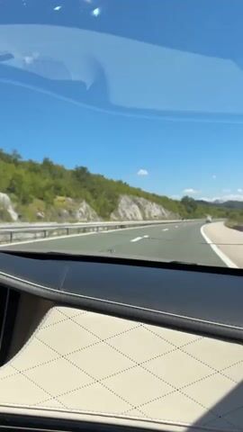 A first-person view from inside a car shows a highway with a clear blue sky and some clouds. The car's dashboard, with its quilted cream and black leather, is visible in the foreground. The road curves to the right, with a guardrail running alongside it. Hills covered in green trees and rocky outcrops are on the left side of the road. Several other vehicles are on the highway in the distance. The sun is shining, indicating it is daytime.