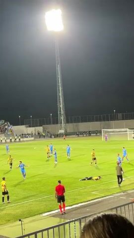 A soccer match is being played at night under bright stadium lights. There are two teams on the field, one wearing blue uniforms and the other wearing yellow and black uniforms. A referee in a red shirt is also on the field. One player in a yellow and black uniform is lying on the ground. The stands are filled with spectators. The field is green grass.