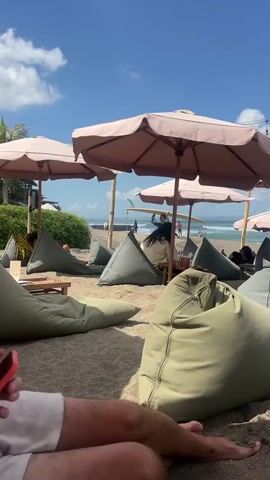 The video shows a beach scene with beanbag chairs and umbrellas on the sand. There are several people visible in the background near the water. One person is walking with a surfboard. The sky is blue with some clouds. The camera is focused on the foreground where a person's legs and hand holding a phone are visible.