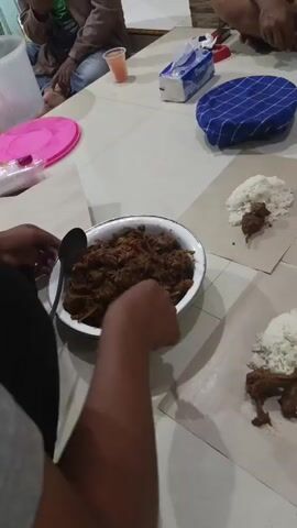 The video shows a group of people eating a meal served on a tiled floor, likely in a casual setting. The food, a brown stew-like dish and white rice, is served on pieces of brown paper arranged on the floor. One person is actively serving themselves from a large white bowl of the stew using a spoon and ladle. Another person's hand is visible reaching for food. In the background, other individuals are seated, and various items like a tissue box, a drink in a plastic cup, and plates are scattered on a table. The lighting appears to be artificial, casting a warm glow on the scene.