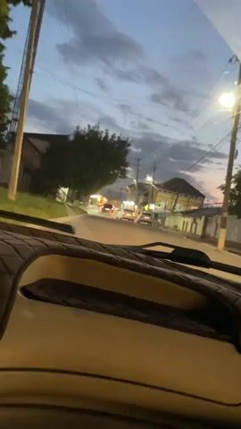 The video, shot from the interior of a vehicle, shows a street scene in Buynaksk, Russia, during dusk. The sky is a deep blue with scattered clouds, indicating the transition from day to night. Streetlights are illuminated, casting a warm glow on the road and surrounding buildings.

Several cars are visible on the road, some with their headlights on, suggesting traffic is moving. The buildings lining the street appear to be residential or small businesses, with a mix of architectural styles. A prominent tree with dark green foliage is on the left side of the frame, partially obscuring a building. Power lines crisscross the sky, a common sight in urban and semi-urban environments.

The interior of the car is partially visible, including the dashboard and a quilted fabric covering, suggesting the camera is positioned behind the windshield. The overall mood is calm and observational, capturing the quiet ambiance of a street at twilight. There is no discernible weather event, and the lighting suggests a clear evening. The time of day is clearly evening, as evidenced by the sky color and artificial lighting. No people are clearly visible, but the presence of cars implies human activity. The focus is on the environment and the drive.