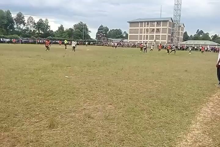 The video depicts a soccer game in progress on a grassy field. There are players wearing both white and orange jerseys. A crowd of spectators lines the edge of the field, and some are standing on a makeshift platform for a better view. In the background, there is a multi-story brick building, a metal tower, and trees. The sky is overcast. Players are running and kicking the ball as the game unfolds.