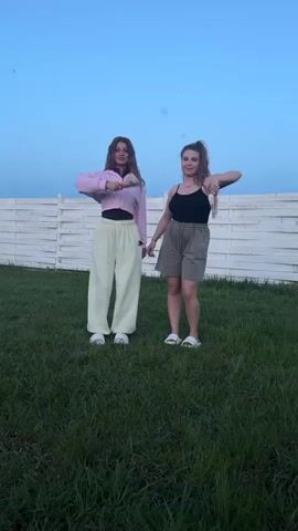 Two young women are standing on a green lawn with a white fence in the background. The sky is a clear blue. The woman on the left is wearing a pink crop top jacket, a black shirt, and loose light yellow pants, along with white sneakers. The woman on the right has on a black tank top, grey shorts, and white sandals. Money is thrown into the air and falls around them. The women raise their arms, smile, and pose for the camera.