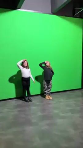 Two young girls stand in front of a green screen in a studio with a gray floor and a black ceiling. Initially, they are a few feet apart, with each girl placing a hand on her head. Then, they cross their arms and turn to face each other. Finally, they stand back to back, still with crossed arms. Throughout, they look toward the camera. The girl on the left wears a white shirt with black leggings and black boots. The girl on the right is wearing a black top with gray camouflage pants and tan boots.