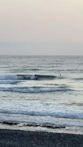 A surfer rides a wave in the ocean. The water is calm, and the sky is overcast. There is a rocky shore in the foreground. The surfer is wearing a dark wetsuit and is standing on a surfboard. The wave is small and appears to be breaking. There is another person in the water in the background, who may also be a surfer. The scene is peaceful and serene.