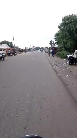 The video pans across a two-lane asphalt road with a shoulder on the right side. The sky is overcast. On the left side of the road, there are buildings, trees, and several motorcycles. Some people are standing near the motorcycles. On the right side of the road, there are more trees and some makeshift stalls or structures. Several people are walking along the shoulder, including one person carrying something on their head and another person in a blue jacket. Multiple motorcycles are seen on the road, some moving and some stationary. There are also some pieces of trash or debris on the road and the shoulder. The overall impression is of a rural or semi-urban setting.