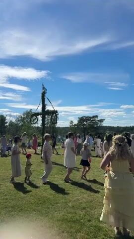 A midsummer celebration is taking place outdoors on a sunny day. A Maypole decorated with greenery and a Swedish flag stands in the center of a grassy field. A group of people of various ages are gathered around the Maypole, some are dancing in a circle while others watch. Children are present, some of whom are dancing. The environment includes a clear blue sky with white clouds, trees in the background, and a body of water visible in the distance. The people are dressed in light, summery clothing.