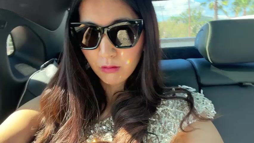 A woman with long brown hair and large, dark sunglasses sits in the back seat of a car. She is wearing a sparkly, sheer top. The car is moving, and the window shows a view of water and a building. The water is sparkling in the sunlight. The sky is clear and blue.