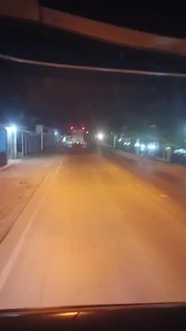 The video shows a view from inside a vehicle driving at night on a road. The road appears to be paved and is illuminated by streetlights and the headlights of the vehicle. There are buildings and trees visible along the sides of the road, and other vehicles can be seen in the distance. The overall scene suggests a relatively quiet nighttime drive through a populated area.