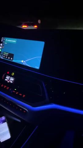 The video shows the interior of a car at night. The dashboard is illuminated with blue ambient lighting. A navigation screen is visible on the left side of the dashboard, displaying a map. Red lights from other vehicles are visible outside the car. The camera is positioned inside the car, looking forward.