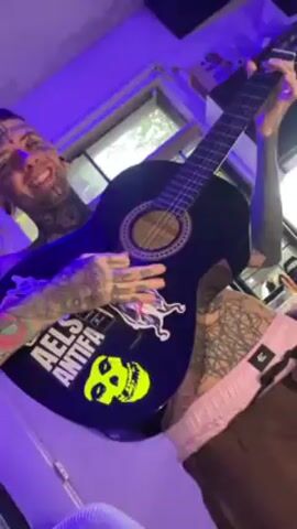 A person with tattoos on their face and body is playing an acoustic guitar with stickers on it. The stickers include "Michaels Antifa" and a yellow skull. The person is smiling and appears to be enjoying themselves. They are in a room with blue lighting, a window, and studio equipment. The guitar has a black finish.