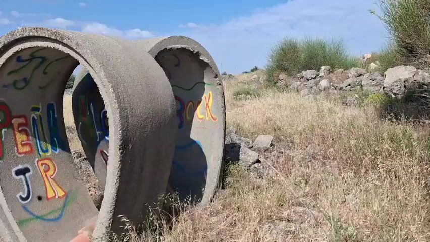 A concrete culvert is shown in a dry, grassy field. The culvert is covered in colorful graffiti. Two orange ceramic pipes are visible inside the culvert. A yellow cartoon face with its tongue sticking out is spray-painted on the inside of the culvert. The camera pans slowly across the culvert and the surrounding landscape.