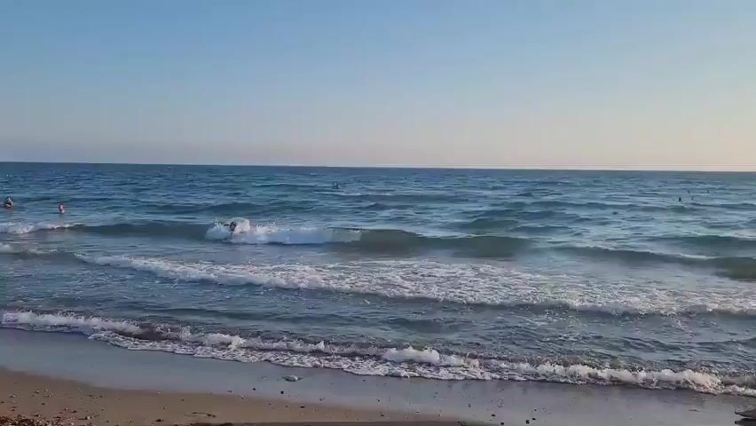 The video shows a man walking along the shore of a beach as waves wash onto the sand. In the distance, several people are swimming in the ocean. The sky is clear and blue, and the sun appears to be setting, casting a warm glow over the scene. The man is wearing dark swim trunks and walks with his back to the camera.