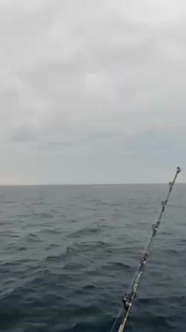 The video shows a fishing rod extending from the bottom right of the frame upwards at an angle, over an open body of water under a cloudy sky. The water is dark and choppy, with small waves visible. The fishing rod is silver and black, with multiple guides along its length. The sky is overcast with gray clouds, and the horizon is a blurred line between the water and the sky.