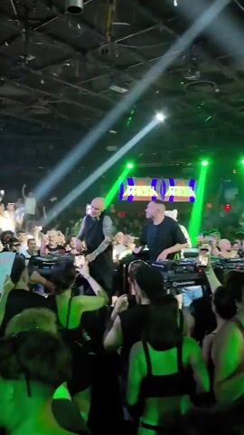The video shows a crowded indoor venue with a DJ setup on a stage. Two male DJs are visible, one on the left with a bald head and tattoos, wearing a dark vest over a dark shirt, and another on the right wearing a dark shirt. They are both standing behind DJ equipment. A large screen behind them displays text that appears to be "SOUL COAST" repeated. Bright green and white spotlights illuminate the stage and the audience. The audience is a dense crowd of people, many of whom are holding up their phones, likely to record the event. The atmosphere appears to be a concert or club event. The camera is positioned within the crowd, looking towards the stage.