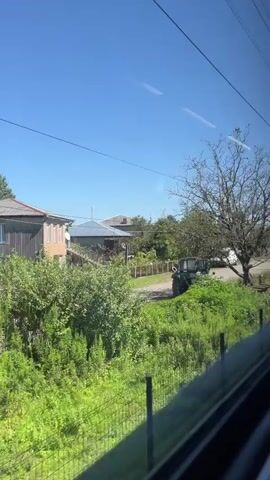 This is a video taken from a moving train. The camera is pointed out the window, showing a rural landscape. In the foreground, a utility pole with the number 260 on it is visible, along with wires and greenery. In the midground, there is a road, grass, and houses. Further in the background, trees and a clear blue sky are visible. The train appears to be moving through a residential area.