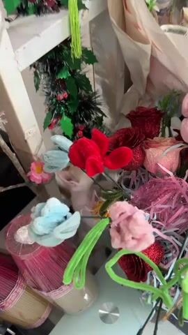 A close-up shot shows a kitten playing with a bouquet of artificial flowers. The kitten's head is visible among the flowers. The bouquet is a mix of red, pink, and blue flowers, with some glittery red roses. A green garland with red berries is also visible in the background. The scene appears to be indoors, with a white surface and white packaging material in the background.