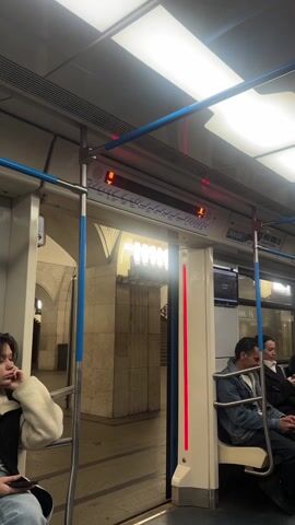 The video shows the inside of a subway car, with a few passengers seated. The door of the subway is open, revealing the station platform. An LED display above the door shows the name of the upcoming station. The subway car is lit by fluorescent lights. A person is leaning against a pole near the door. Other people are sitting down, looking at their phones or just observing their environment. The station platform is made of marble. There are escalators visible, leading to the station entrance. The overall ambiance suggests a routine subway ride.