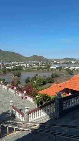 This is a vertical video filmed from a high vantage point looking out over a town and surrounding landscape. In the foreground, there is a paved patio area with a low stone wall adorned with white balusters and red flowers. A set of stone stairs with metal railings is visible leading down. Beyond the patio, a town with buildings and some flooded rice paddies can be seen. In the distance, rolling green hills under a clear blue sky form the backdrop. Several people are walking on the patio. The lighting suggests it is daytime, likely midday, with strong sunlight casting distinct shadows.