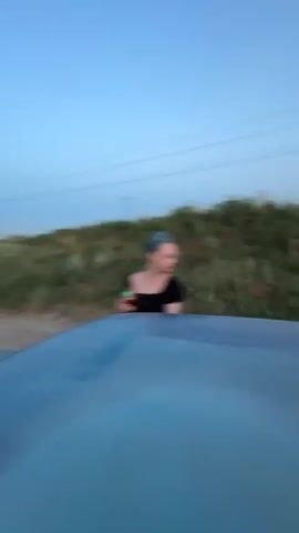 The video captures a car ride through a rural area in Milyanfan, Kyrgyzstan. The time of day appears to be late afternoon or early evening, with the sun low in the sky, casting a warm glow and creating a hazy, bright atmosphere. The landscape is characterized by a dirt road winding through a grassy, somewhat dry terrain, suggesting a rural or semi-arid environment.

The primary subject is a young woman with bright red hair, leaning out of the passenger window of a moving vehicle. She is wearing a light-colored t-shirt with a graphic design that includes a character resembling Hello Kitty and the text "Love" and "Happy" in pink. She also appears to be wearing pink shorts. Her expression is relaxed and perhaps a little playful as she looks towards the camera. Her arm is resting on the open car door, and there's a glimpse of a tattoo on her forearm.

The video is shot from a low angle, possibly from another vehicle or from within the same car, looking towards the woman and the scenery. The motion blur is significant, indicating movement and adding a dynamic, spontaneous feel to the footage. The overall mood is one of freedom and enjoyment, possibly capturing a moment of roadside adventure during a trip. The natural light and open road contribute to a sense of casual exploration and a connection with the surroundings.