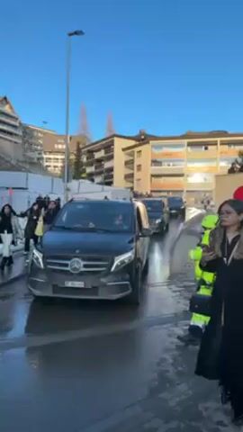 The video shows a street in front of a building with a large Mastercard sign. There is a group of people gathered near the sign, some of whom appear to be photographers or media personnel, with cameras and equipment. Several cars are parked on the street, including a black Mercedes van. A person in a bright yellow vest is directing traffic. Other people are walking on the street. The weather appears to be sunny and the sky is clear. There are buildings in the background and a tree to the right of the Mastercard sign.