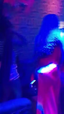 The video shows two women dancing in a room with colorful lighting. One woman is wearing a striped top and shorts, while the other is wearing a red dress. The room has a shiny textured wall. They appear to be enjoying themselves.