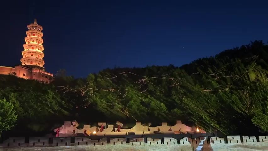 At night, a tall pagoda on a hill shines with bright spotlights against a dark blue sky. The hill is covered in lush green trees. Below the hill, a decorative wall with crenellations and figurines stands illuminated. There are no people visible, and the scene is still.