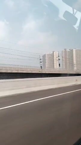 The video captures a view from a moving vehicle, likely a car or bus, traveling on a highway. The camera is angled to show a perspective from inside the vehicle looking outwards. In the foreground, a portion of the highway asphalt is visible, marked with white lines, indicating lanes of travel. Beyond the highway, a concrete barrier separates the road from an elevated railway track. The railway track is supported by pylons and overhead electrical wires for a train. In the background, several tall, multi-story apartment buildings are visible. These buildings appear to be modern and residential. The sky is overcast with a light haze. The overall scene depicts an urban or suburban environment, likely in a city with a developed transportation infrastructure.