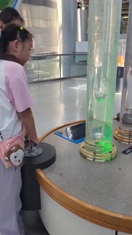 Two young girls are at a science museum exhibit. The girl on the left, wearing glasses and a white shirt, stands with her head tilted slightly, observing. The girl on the right, wearing glasses and a pink and white shirt, is leaning forward and interacting with a circular mechanism on a pedestal. She has a pink bag with a cartoon character design slung over her shoulder. In front of them is a tall, clear cylindrical tube filled with water and several large bubbles. The tube is illuminated with green light from the bottom, causing the bubbles to glow. The exhibit appears to be part of a larger indoor space with polished floors and elevated walkways visible in the background.