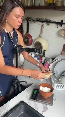 A woman with long brown hair wears a dark blue dress and a white necklace. She is standing in a kitchen, with her left hand holding a small bag of something, pouring it into a wooden bowl. Her right hand is holding a spoon, and she is scooping some of the contents from the bag into the bowl. The bowl is sitting on a digital scale. There is a tripod with a phone attached behind her. In the background, there are shelves with jars and bottles, pots hanging on the wall, and a dish rack with plates and bowls. The counter is white and marbled.
