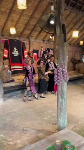 Three people are standing inside a rustic wooden structure with a thatched roof. The person on the left is wearing traditional clothing with intricate patterns and a headdress. The person in the middle is wearing a floral-patterned top and a black skirt. The person on the right is wearing a traditional outfit with black and pink accents. They are all smiling and appear to be dancing or posing for a photo. Behind them, traditional clothing is displayed on the walls. In the foreground, a bunch of red chilies and garlic are hanging from a wooden post.