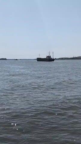 A barge is seen in a wide river under a clear sky. The barge is moving from left to right across the frame. The water is choppy with sunlight glinting off the surface. In the background, a distant shore is visible with low hills and some man-made structures, possibly towers or pylons. The scene is calm and suggests a daytime environment.
