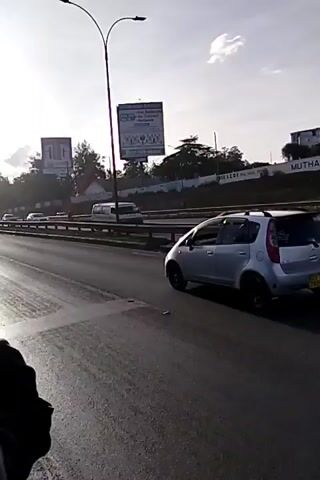 The video shows a silver hatchback car driving along a highway. There are other cars visible on the highway and in the opposite lane. Several billboards can be seen alongside the highway. The sky is clear with a few clouds.  The video is recorded from a moving vehicle.  A portion of the person holding the camera is visible on the left side of the frame. The silver car has black wheels and a roof rack. The license plate is partially visible.