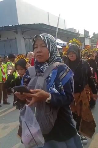 The video shows a group of young women wearing black hijabs and patterned skirts participating in what appears to be a parade or procession. They are walking outdoors, possibly on a street lined with buildings. Some of the women are smiling and interacting with each other, while others are focused ahead. One woman in the foreground is holding a green object, possibly a megaphone or a toy. The overall mood seems festive and celebratory.