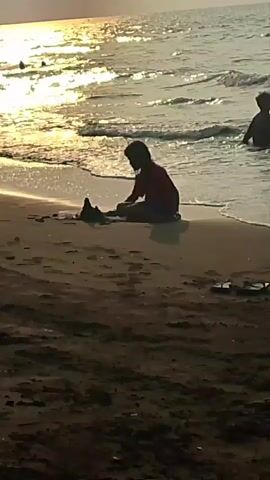 A medium-shot video shows a sandy beach with the ocean in the background. The sun is setting, casting a golden reflection on the water. Two children are visible in the scene. In the foreground, a child wearing a red shirt is sitting on the sand, playing with a sandcastle. Further back, in the water, another child is standing, seemingly playing or swimming. The overall atmosphere is peaceful and idyllic.