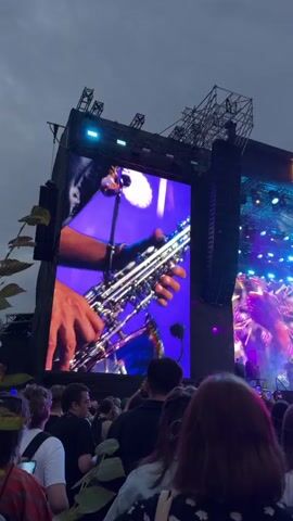 The video shows a crowded outdoor music festival. A large screen displays a close-up of a person playing a saxophone, with bright purple and blue lights in the background. Above the screen, the words "PARK LIVE" are visible in illuminated letters. The crowd in the foreground consists of people, many of whom are holding up their phones to record the performance. The sky is overcast.