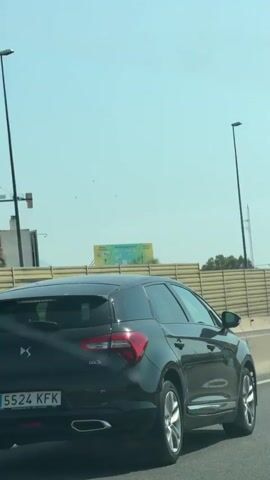 The video shows the rear view of a black DS 5 car driving on a highway. The car has a license plate reading "5524 KFK". The scene is a sunny day with a clear blue sky, suggesting it is daytime. The car is driving on the right side of the road, with other vehicles visible in the background.

In the distance, there are signs for "Carrefour San Juan", indicating a commercial area. There are also traffic lights and overhead streetlights lining the highway. A brown fence runs along the side of the road, and beyond that, there are hills with sparse vegetation and some buildings. The overall setting appears to be an urban or suburban highway in Alicante, Spain.

The activity is simply driving on the highway. There are no people clearly visible inside the car or interacting with it. The weather is bright and sunny. The overall mood is neutral, depicting a typical scene of road travel.