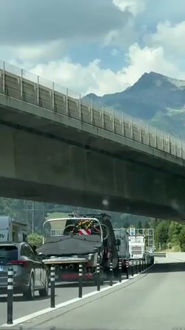 The video shows a traffic jam on a highway with a large concrete overpass above. The camera is inside a vehicle looking forward. On the right side of the road, there is a line of cars, RVs, and campers. Some of the vehicles have bicycles mounted on top. The road is lined with black and white bollards. In the background, there are green, tree-covered mountains and a peak with snow on top. The sky is blue with some white clouds. The traffic appears to be moving slowly or stopped. There are signs and traffic lights visible in the distance.