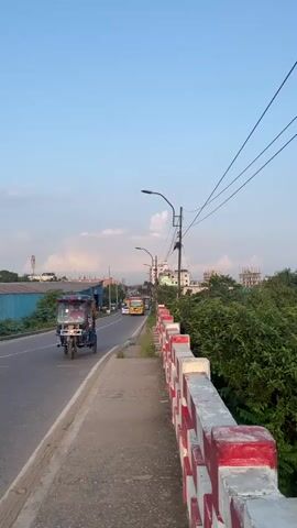 The video shows a clear blue sky with some scattered clouds above a road with a sidewalk. On the left side of the road, there is a blue building, a white car, a yellow bus, and a blue and red motorized rickshaw. On the right side of the sidewalk, there is a red and white striped railing. There are green trees and some buildings in the background. The video appears to be taken during the daytime.