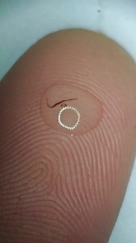 A video shows a close-up of a finger with a drop of liquid on it. A dark, thin, curved object is visible in the liquid. The next shot shows the hand holding the finger with the droplet, under a magnifying lamp. The lamp is white and has the word "MAGUS" on it. The background is dark and contains other objects.