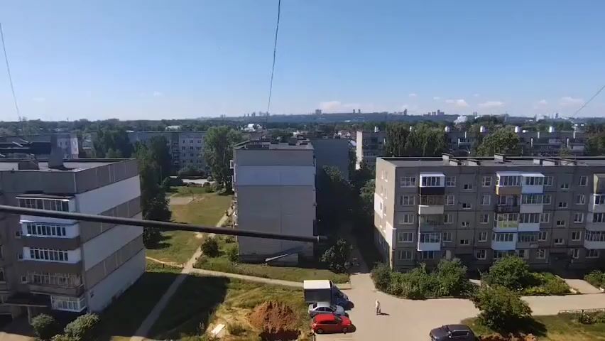 This is a video footage capturing a view from a high vantage point overlooking a residential area in Bor, Russia. The scene is dominated by several multi-story apartment buildings, typical of Soviet-era architecture, with grey facades and numerous balconies. In the foreground, parts of at least two apartment blocks are visible, one on the left and another on the right. In the middle ground, more apartment buildings are interspersed with trees, creating a dense urban landscape. Beyond these, a distant city skyline can be seen on the horizon, with numerous taller buildings indicating a larger urban center. The sky is clear and blue, suggesting a sunny day, likely during the daytime, given the strong shadows and bright illumination.

The ground level shows some patches of dry grass and dirt, along with a few parked cars. One person is visible walking in the open space between buildings, adding a human element to the scene. There are also piles of reddish-brown material, possibly soil or construction debris, near the buildings. The overall impression is of a typical day in a residential district, characterized by functional architecture and a sprawling urban environment. No text is visible within the video frames. The weather appears to be warm and clear, conducive to outdoor activities, though the scene itself is tranquil, with no obvious signs of immediate action or events. The high angle suggests the footage was taken from a drone, a tall building, or a crane.