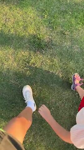 This is a first-person perspective video clip that begins with a close-up shot of someone's feet and legs walking on a grassy lawn. A child's feet, wearing pink sandals and red shorts, are visible next to the person's feet. The camera then pans up and to the right, revealing a scenic park-like setting.

The scene is a well-maintained park with lush green grass. In the background, there is a charming small suspension bridge with wooden planks and railings, decorated with some sort of rustic attraction, possibly a water wheel, to its left. Beyond the bridge, there are various trees, including a prominent weeping willow with its branches hanging low, and other leafy trees. In the far distance, mountains rise against a clear blue sky. The sunlight suggests it is either early morning or late afternoon, casting soft shadows on the grass. The overall atmosphere appears peaceful and pleasant, suggesting a leisurely stroll in an outdoor recreational area. No text is visible in the clip.