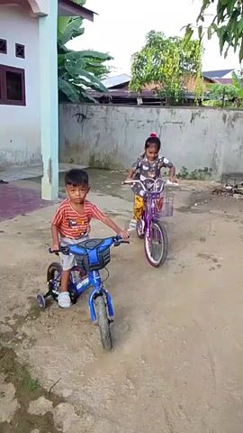Two young children, a boy and a girl, are riding bicycles in a dusty, outdoor area. The boy, wearing an orange and white striped t-shirt and shorts, is on a blue bicycle with training wheels. The girl, in a grey t-shirt with a cartoon bear pattern and yellow, patterned pants, is on a purple bicycle, also with training wheels. Both children appear to be concentrating as they pedal. The girl is smiling and looking ahead, while the boy looks down at his bicycle.

The setting appears to be a residential area in Langsa, Indonesia. In the background, there's a concrete wall, a building with a light blue pillar, and lush green vegetation, including banana plants. The ground is dirt and appears a bit damp in places. The lighting suggests it is daytime, possibly late morning or early afternoon, with no strong shadows. The overall mood is one of innocent childhood play. There is no visible text in the video.