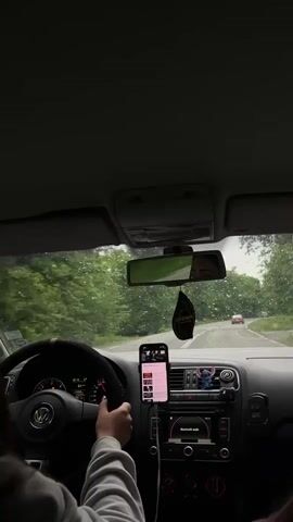 The video shows a person driving a car on a rainy day. The person's left hand is on the steering wheel, and their phone is mounted in the center console, displaying a music playlist. Raindrops are visible on the windshield. The car is driving on a road surrounded by green trees. The car's dashboard and interior are visible, including a Stitch figure on the dashboard and an air freshener hanging from the rearview mirror. A car is visible in the distance. The person is wearing a light-colored sweatshirt. The car is a Volkswagen Polo. The car radio is playing music.