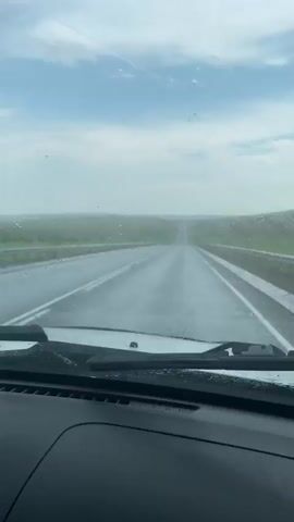 The video shows a view from inside a car driving on a wet road, likely in the rain, as indicated by water droplets on the windshield and the appearance of the road surface. A windshield wiper is visible in the foreground, at times clearing the view. The road ahead is empty and curves slightly, disappearing into the distance. The landscape on either side of the road is green and appears to be a flat or gently rolling terrain, with a cloudy sky above. The overall impression is one of driving through a somewhat overcast or rainy day.
