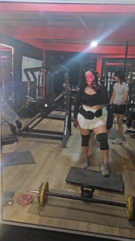 The video shows a woman in a gym taking a selfie. She is standing on a black mat, wearing a black top, a light-colored skirt, knee pads, and gray sneakers. She has curly hair and is holding a pink phone in her left hand. In front of her is a barbell with yellow weights on each side. In the background, there is gym equipment, including weight machines and racks. Other people are present in the gym, including another woman walking behind the main subject. The gym has a red ceiling and wooden floors.