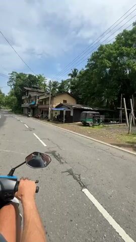 The video shows a first-person perspective of someone riding a blue scooter down a paved road. The road has a white dividing line and a shoulder. On the left side of the road are buildings, including one with a shop called "RUCH Communication" and "Book Shop." There are parked motorcycles in front of the buildings. The surroundings are green with trees and foliage. The sky is partially cloudy. The rider's arm is visible in the foreground, holding the scooter's handlebar. The video appears to be taken during the day.