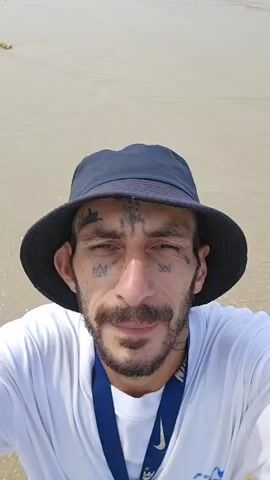 A man with face tattoos, a beard, and a mustache is featured in a selfie video on a sunny beach in Cam Ranh Ward, Vietnam. He is wearing a dark bucket hat, a white long-sleeved t-shirt, and a blue lanyard with the "NIKE" logo. The t-shirt also has text that appears to say "Hang Loose." A watch is visible on his left wrist. He smiles throughout the video and waves to the camera at 0:03. The background shows a sandy beach, clear turquoise ocean water with gentle waves, and a bright blue sky with scattered white clouds, indicating pleasant daytime weather.