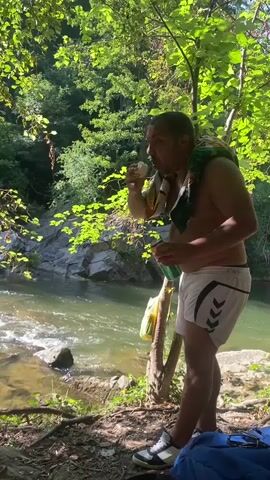 The video shows a man standing by a river with a towel draped over his shoulders. He is wearing white shorts and sneakers. He is holding a green bottle and a yellow object in his hands. In the background, there is a rocky riverbank with trees and foliage. Another person is partially visible in the river in the distance. There are bags and towels on the ground near a large rock. The sun is shining through the trees, creating dappled light on the scene. The man turns his head to the left as the video continues, and then the camera pans to the right, showing more of the river and the surrounding woods.