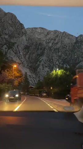 A video taken from inside a vehicle looking out at a winding road at dusk. The road has white lane markings and is illuminated by streetlights. On the left side of the road, there are trees and a mountain in the background. On the right side of the road, there are more trees, a stone wall, and a building. The sky is a deep blue with a few clouds. The overall impression is of a scenic drive through a mountainous area as night approaches.